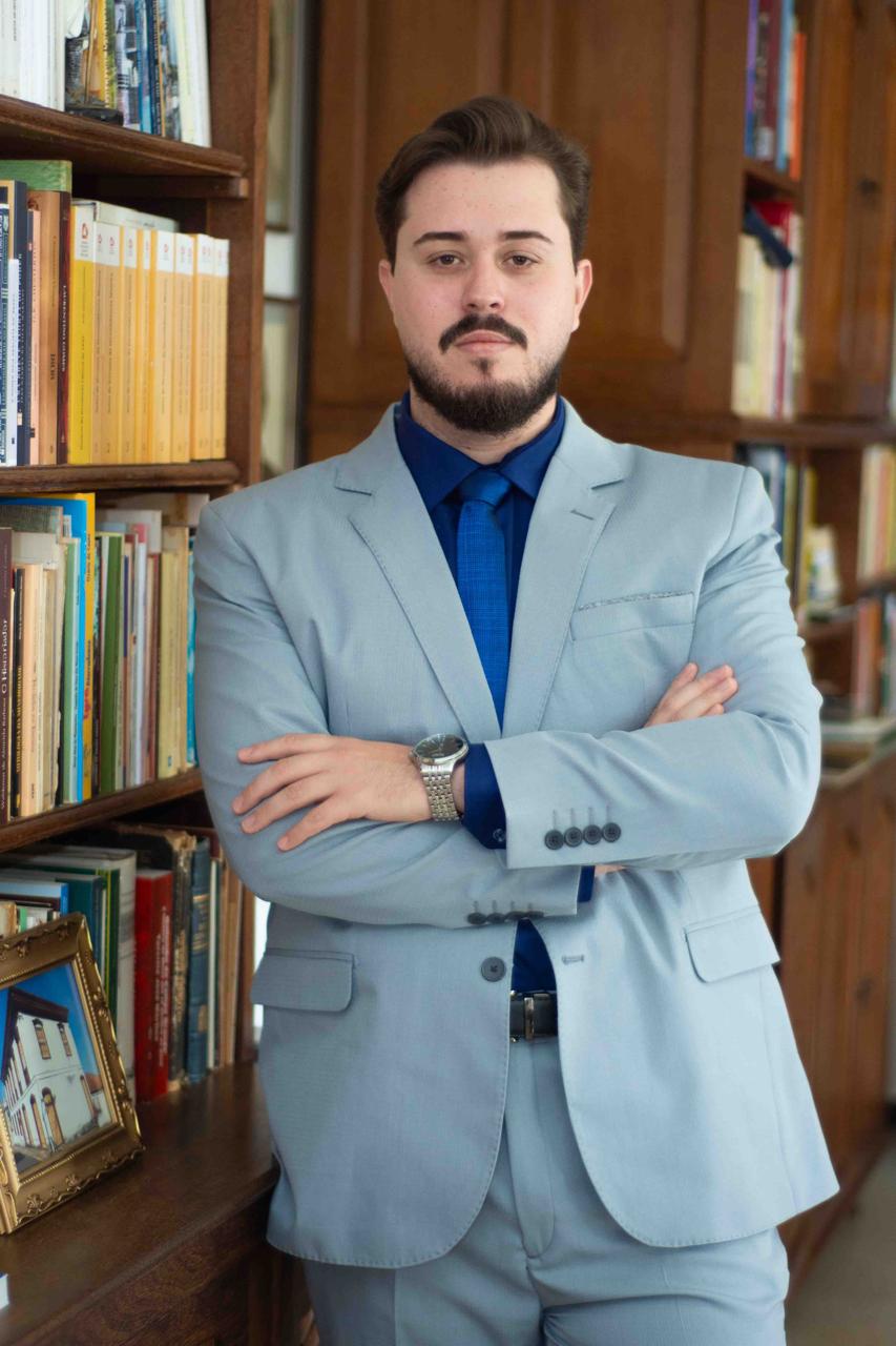 Matheus Campos sentado em seu escritório, entre estantes de livros jurídicos